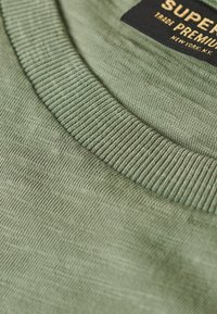 T-shirt verde in cotone con colletto tondo a costine, texture morbida e un'etichetta con "Super Premium" in oro su sfondo nero.