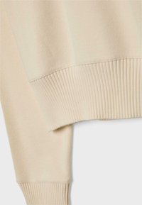 Manche de pull beige en tricot et détail de bord-côte sur un fond clair, montrant en gros plan la texture et le tissage du tissu.