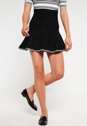 Femme portant une jupe évasée noire avec un galon zigzag blanc, un haut rayé et des mocassins noirs, posant avec une jambe croisée sur l'autre.