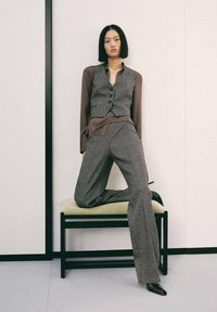Chaleco gris ajustado sobre una camisa de botones marrón, combinado con pantalones anchos grises. Texturas suaves con un diseño minimalista. Tacones altos negros.