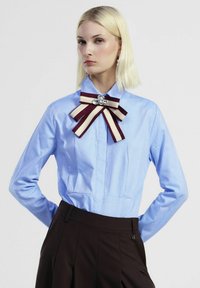 Blusa azzurro chiaro con colletto, con fronte plissettato, caratterizzata da un fiocco a righe bordeaux e crema e da un accento a gemma sullo scollo.
