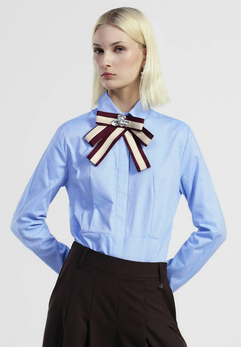 Blusa azzurro chiaro con colletto, con fronte plissettato, caratterizzata da un fiocco a righe bordeaux e crema e da un accento a gemma sullo scollo.