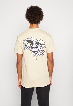 Camiseta de algodón beige con un diseño gráfico en la espalda que muestra un rostro y manos rompiendo el fondo, delineados en negro y acentuados con ojos amarillos.
