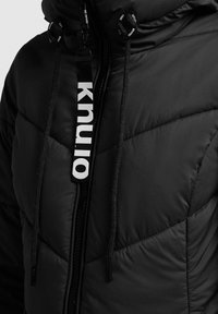 Chaqueta acolchada negra con diseño de rombos, que cuenta con cierre de cremallera, cordones ajustables y una etiqueta de marca con "KNU."