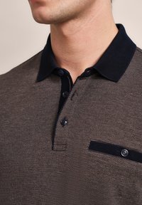 Texturiertes braunes Poloshirt mit navyfarbenem Kragen, zwei Knöpfen und einer Brusttasche mit einem dunklen navyfarbigen Akzent. Nahaufnahme des Ausschnitts.