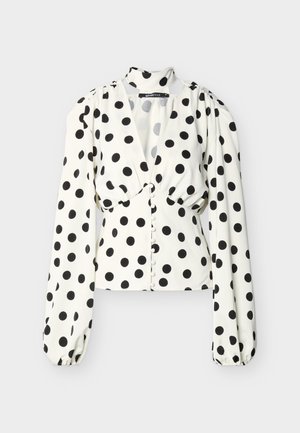 Λευκό πουκάμισο με μαύρες πουά βούλες, μακριά φουσκωτά μανίκια, βαθύ V-λαιμόκοψη και κουμπιά καλυμμένα με ύφασμα κατά μήκος του μπροστινού μέρους.