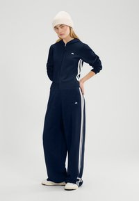 Donna in piedi con le mani dietro la schiena che indossa una felpa con cappuccio blu navy con cerniera, pantaloni a gamba larga con strisce bianche, berretto lavorato a maglia color crema e scarpe da ginnastica bianche.