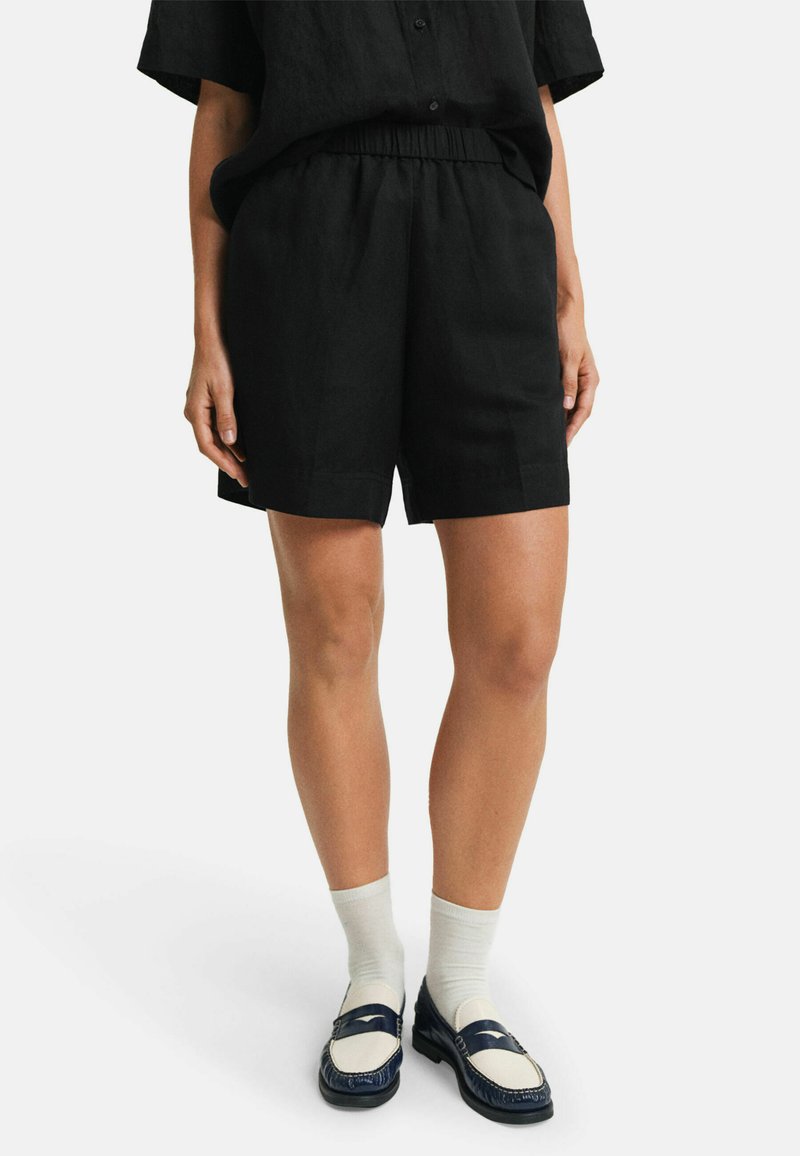 Zwarte linnen shorts met een elastische tailleband, met een losse pasvorm en zijsplitten voor bewegingsgemak. Gecombineerd met marineblauwe instappers en sokken.