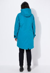 Turquoise waterdichte parka met een capuchon, met een rechte snit, lange mouwen en verstelbare manchetten, gecombineerd met zwarte broek en laarzen.