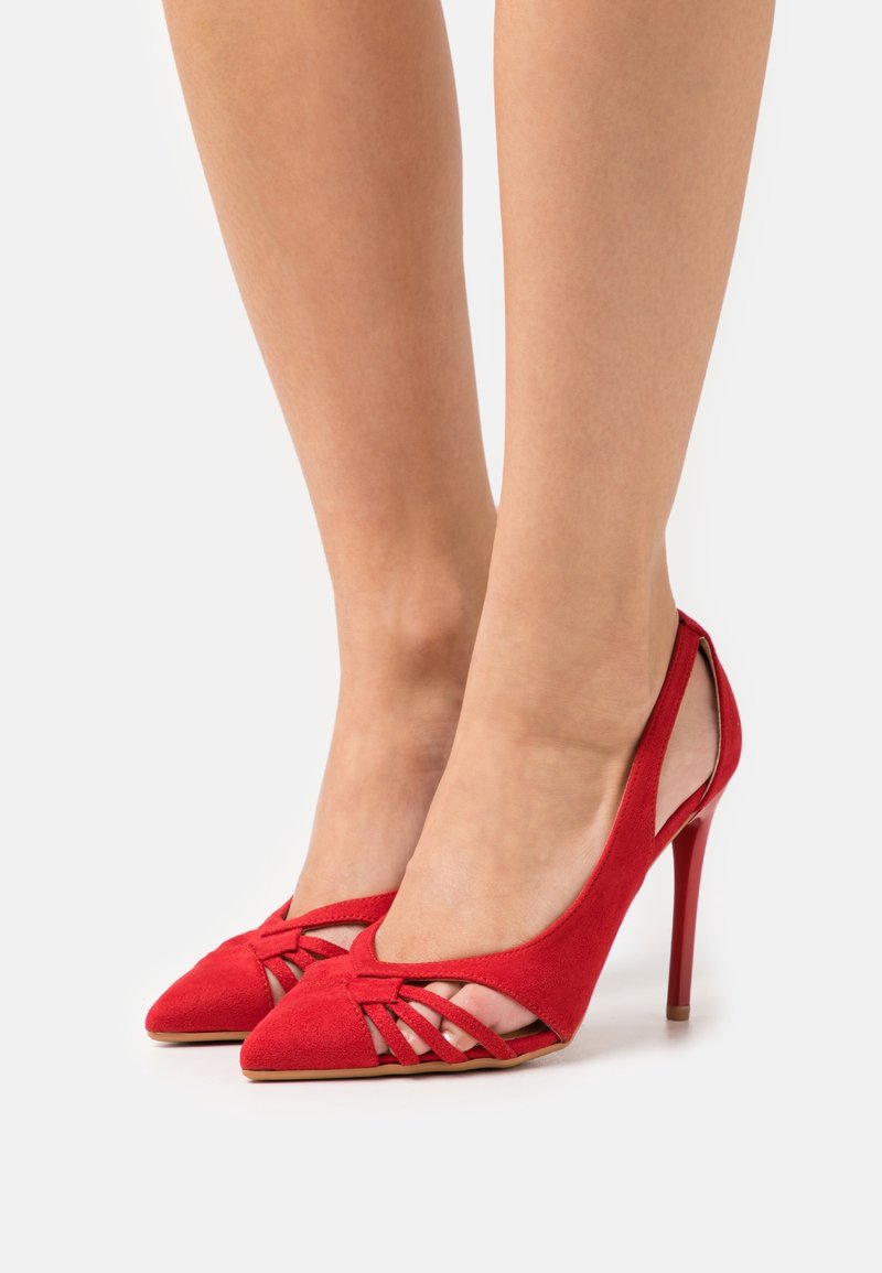 Jalat yang mengenakan sepatu hak tinggi merah dengan sisi yang terbuka dan detail tali anyaman, ditampilkan di latar belakang putih polos.