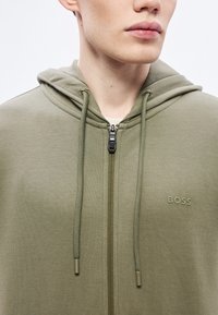 Felpa con cappuccio e zip in verde oliva, realizzata in tessuto morbido. Presenta una zip frontale, coulisse e un marchio in rilievo discreto.