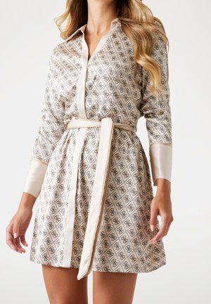 Frau trägt ein cremefarbenes Satin-Kurzkleid mit einem wiederholten schwarzen Logomuster, langen Ärmeln, breiten Manschetten und einem passenden Gürtel, der an der Taille gebunden ist.