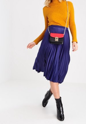 Kobieta w musztardowym golfie, plisowanej granatowej spódnicy midi, czarnych botkach na kostce oraz torebce typu crossbody z czerwoną klapą i łańcuszkowym paskiem.