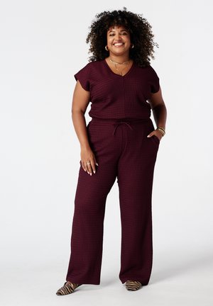 Femme souriante aux cheveux bouclés, debout, portant un haut texturé bordeaux à manches courtes et un pantalon, des chaussures à rayures de tigre et des bijoux en or.