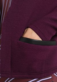 Lila texturerad tyg med en sidficka som har svart läderkant. Designen visar ett lager av plagg med randiga detaljer under.