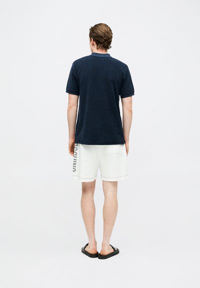 Άνδρας που στέκεται γυρισμένος προς τα πίσω φορώντας μπλε κοντομάνικο πόλο, άσπρες βερμούδες με κείμενο στο πλάι και μαύρες σαγιονάρες.