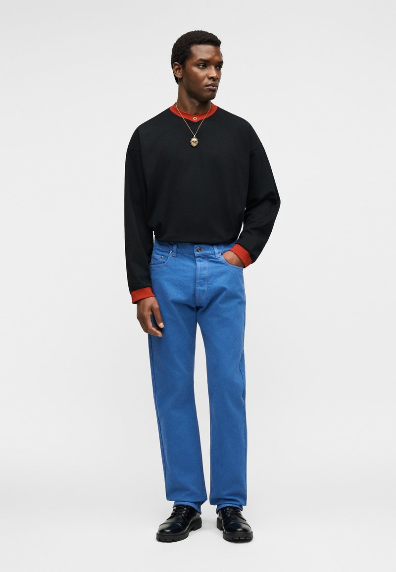 Homme debout, main gauche dans la poche, portant un pull noir avec des poignets rouges, un jean bleu, des chaussures noires et un collier avec un pendentif.