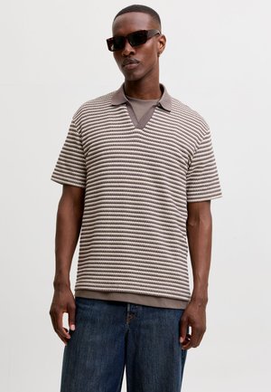 Homme portant des lunettes de soleil foncées, un pull en maille rayé à manches courtes par-dessus une chemise marron à col, et un jean bleu, debout devant un fond blanc uni.