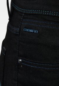 Sort denim med kontrasterende blå syninger, prydet med et G-Star-logo og en sølvknap. Teksturen er glat med en let glans.