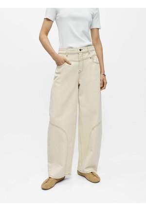 Femme portant un pantalon beige taille haute à jambes larges avec des détails de couture, un haut blanc à manches courtes et des chaussures plates marron clair, debout avec une main dans la poche.