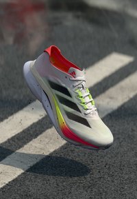 Letvægts sportssko med en hvid mesh-overdel med sorte striber, lyserøde og gule detaljer samt en polstret sål.