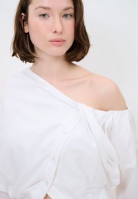 Blouse blanche à épaules dénudées avec fermeture à boutons, en tissu doux, présentant un design drapé et une coupe décontractée, mettant en valeur la zone de la clavicule.