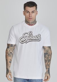 Balts kokvilnas T-krekls ar īsām piedurknēm, kurā ir izteikts melns Siksilk logo kursīvā. Krekls ir ar apaļu izgriezumu un brīvu piegriezumu.