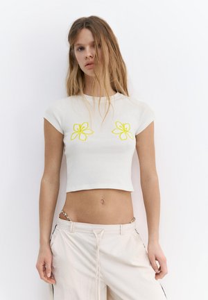 Giovane donna con lunghi capelli biondi indossa una maglietta corta bianca con disegni di fiori gialli e pantaloni bianchi larghi su uno sfondo bianco uniforme.