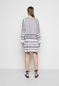 Femme aux longs cheveux portant une robe imprimée noire, blanche et grise jusqu'aux genoux et des sandales noires à lacets, se tenant devant un mur gris uni.