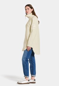 Beige fleece pullover med høj krave, sidelukning og et logo patch på venstre ærme; parret med opsmøgede blå jeans og hvide sneakers.