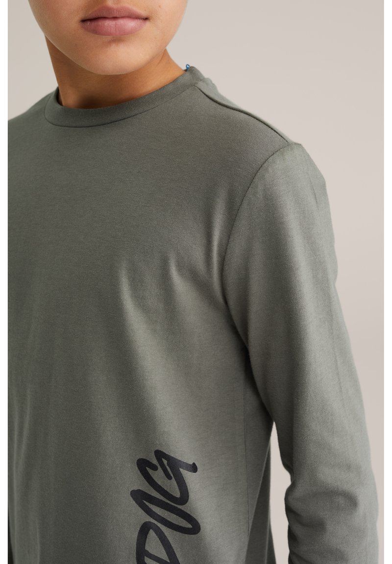 Hosszú ujjú ing sötétzöld anyagból, sima textúrával. A bal alsó oldalon fekete grafikai dizájn található, valamint kerek nyakkivágása van.