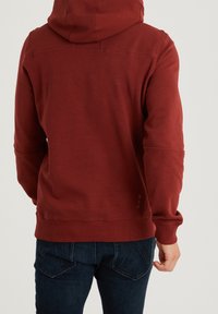 Sudadera burdeos con capucha, de tejido texturizado, puños y dobladillo acanalados. Presenta acentos sutiles en la parte trasera, combinada con jeans oscuros.