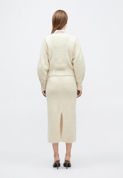 Lõngast kootud kreemjas pullover mahukate varrukatega ja soonikkoes äärega, kombineeritud keskmise pikkusega seelikuga, millel on ees lõhik lisadetailina.