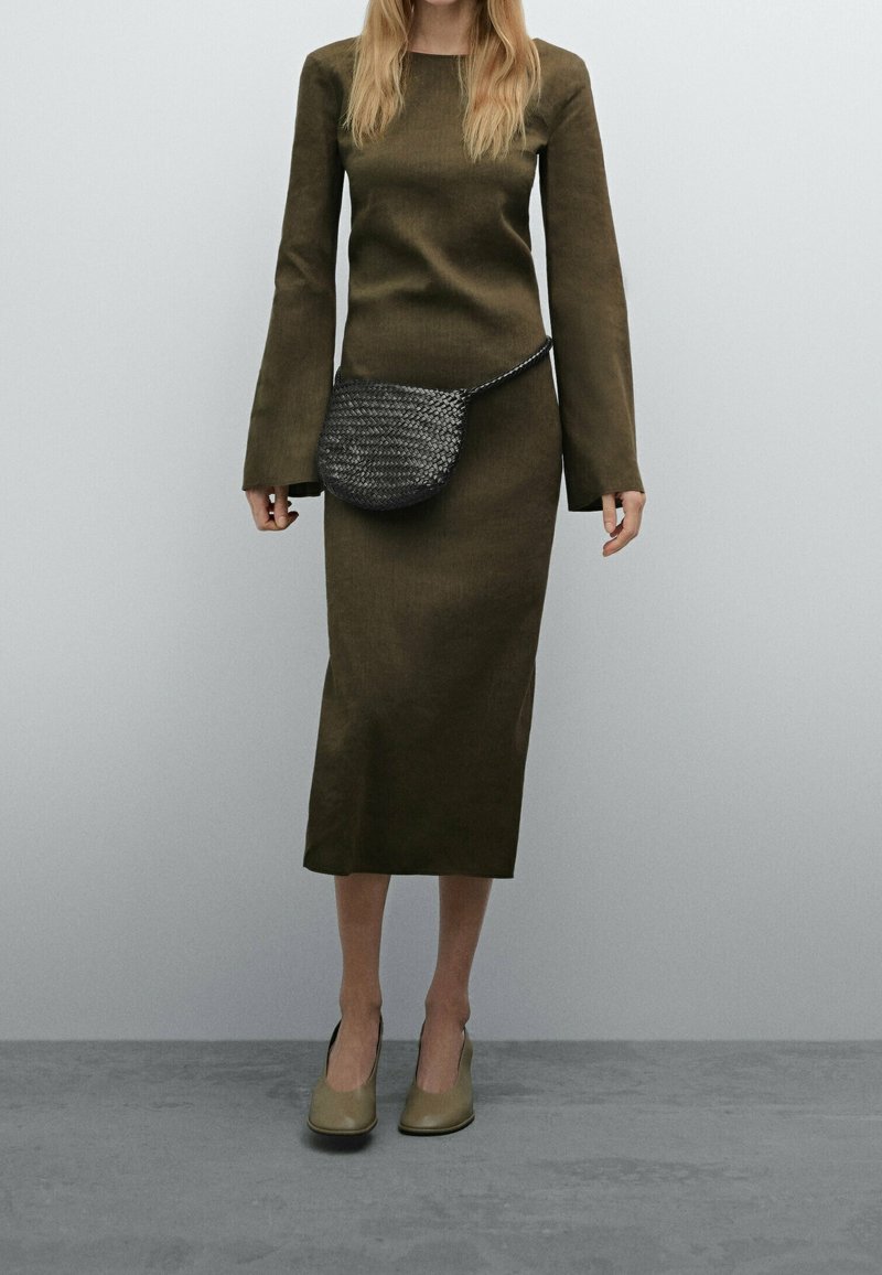 Femme portant une robe midi kaki à manches longues avec un sac ceinture tressé noir et des chaussures à talons beiges, debout sur un sol gris.