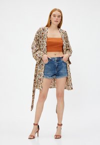 Kimono w kwiatowy wzór w odcieniu beżowym, zestawiony z pomarańczową bluzką bez ramiączek i niebieskimi szortami dżinsowymi. Brązowe sandały na obcasie dopełniają stylizację.