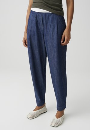 Person, die dunkelblaue lockere Hose, olivgrünes Oberteil und weiße gewebte Slip-on-Schuhe trägt und vor einem hellgrauen Hintergrund steht.