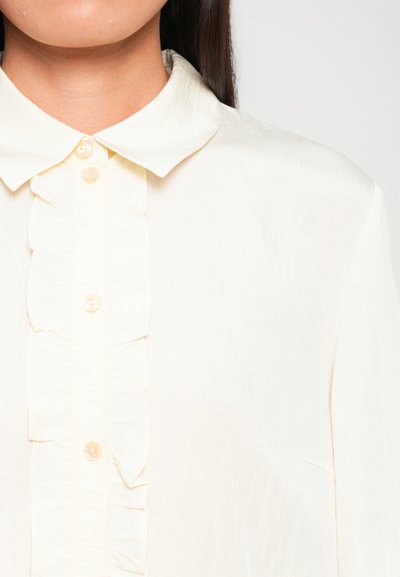 Kreemiline pluus pehme tekstuuriga, millel on krae ja ees ripsmete detail. Ülesse on kinnitatud heledate nuppe püstpinnal.