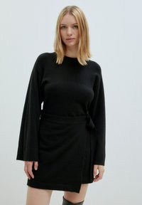 Schwarzes Strickkleid mit langen Ärmeln, wickelndem Design und gebundenem Taillenbund. Weicher Stoff und knielange Länge mit geradem Saum.