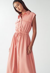 Koralo rožinė suknelė su trumpomis rankovėmis, dviem krūtinės kišenėmis ir elastine juosmens raišteliu. Lygus audinys su laisvai krintančia A-formos siluetu.