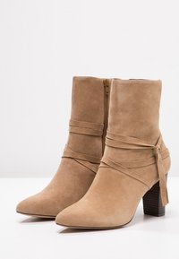 Botines de ante marrón claro con punta afilada, tacón apilado y correas decorativas envueltas. Cuenta con una cremallera lateral para el cierre.