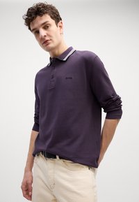 Πουκάμισο πόλο με μακριά μανίκια σε σκούρο μωβ χρώμα, με αντίθετο λευκό περιποιημένο γιακά, διαθ�έτει τρία κουμπιά και κεντημένο λογότυπο στο στήθος.