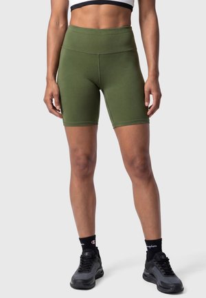 Kobieta ubrana w oliwkowe, wysokowalne spodenki rowerowe, czarne skarpety Champion i czarne sneakersy, stojąca na jednolitym tle.