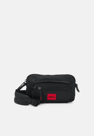 Czarna prostokątna torebka na ramię z regulowanym paskiem, przednią kieszenią na zamek i czerwoną naszywką z logo "HUGO" na przedniej kieszeni.