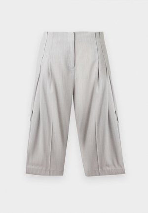 Femme portant une chemise blanche boutonnée avec manches retroussées, un short ample gris clair arrivant au genou, et des talons blancs pointus avec un petit talon.