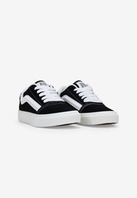 Zapatillas de ante negras con cordones blancos, tira lateral blanca y suela de goma plana. Cuenta con un collar acolchado y detalles de costura.