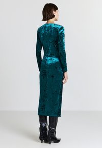 Robe en velours teal avec un design ajusté, des manches longues et une longueur jusqu'aux genoux. Portée avec des bottes noires jusqu'aux genoux. Texture lisse et riche.
