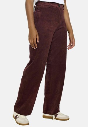 Person wearing wide-leg dark brown corduroy pants and white sneakers with gum soles, standing against a plain light background.