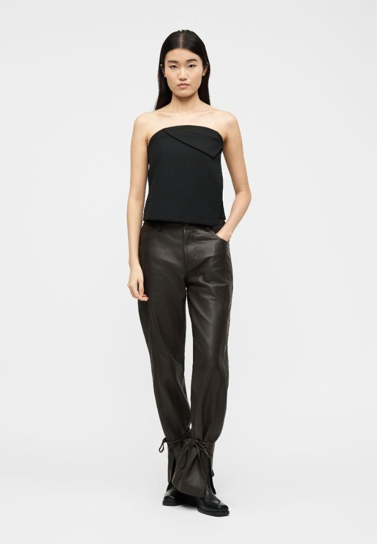 Femme debout portant un haut noir sans bretelles, un pantalon en cuir marron foncé noué aux chevilles, et des bottes noires, sur un fond blanc uni.
