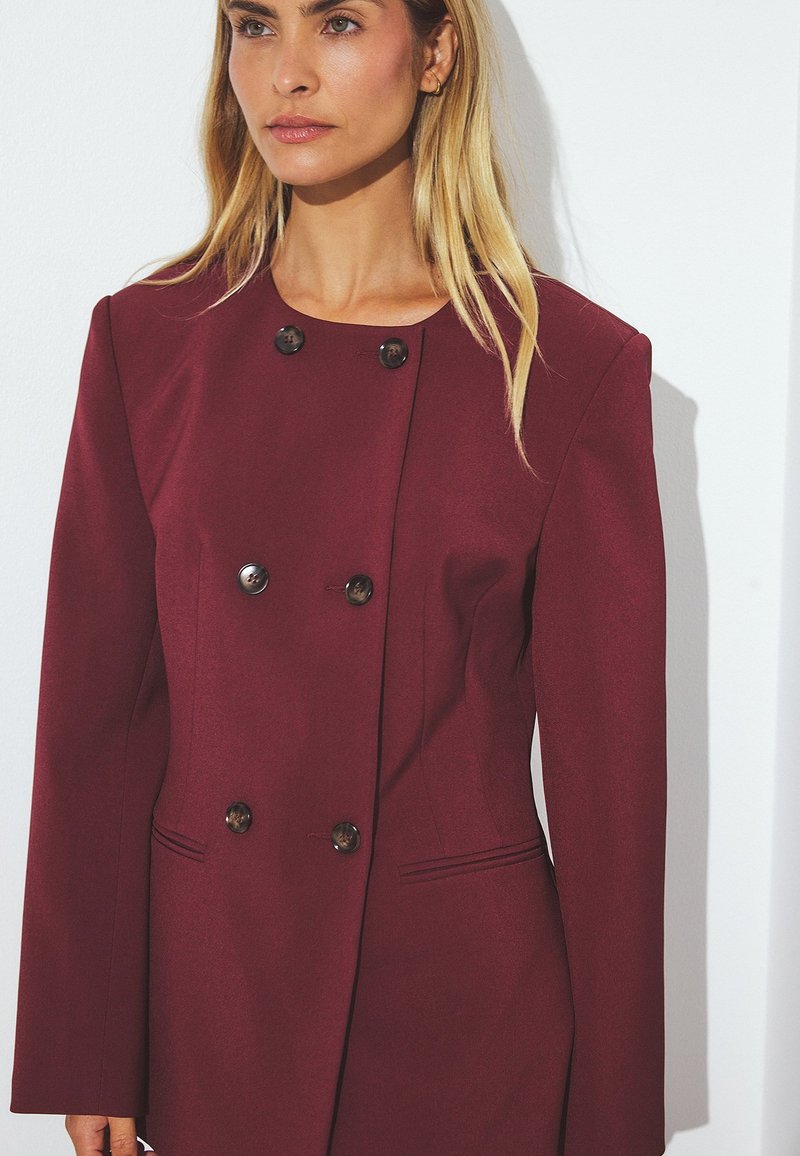 Burgundi struktūruotas švarkas su dvigubo mygtuko uždarymu, ilgomis rankovėmis ir kampuotomis šoninėmis kišenėmis. Lygus audinys, minimalus detalizavimas.