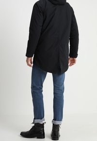 Parka noire isolante avec capuche, présentant un ourlet arrière plus long. Associée à un jean en denim bleu et des bottes noires à lacets.